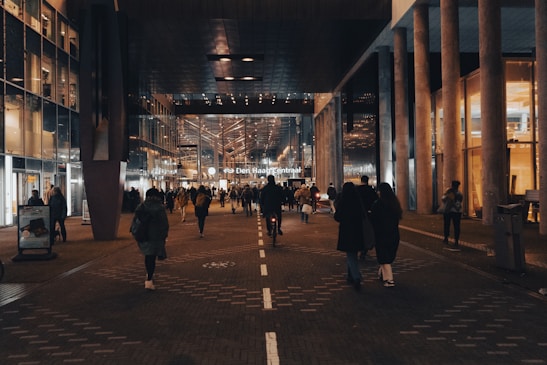 A bustling urban scene featuring a crowd of people walking through a well-lit corridor surrounded by modern architecture. Glass and metal structures are prominent, reflecting the ambient lights. There's a central train station at the end of the walkway, indicating public transport connectivity.