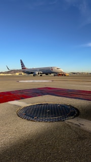An airplane on the tarmac at an airport with the American Airlines logo. The sky is clear and blue, and there are distant hills and airport structures in the background. The foreground shows a textured manhole cover and painted markings on the ground.