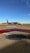 An airplane on the tarmac at an airport with the American Airlines logo. The sky is clear and blue, and there are distant hills and airport structures in the background. The foreground shows a textured manhole cover and painted markings on the ground.
