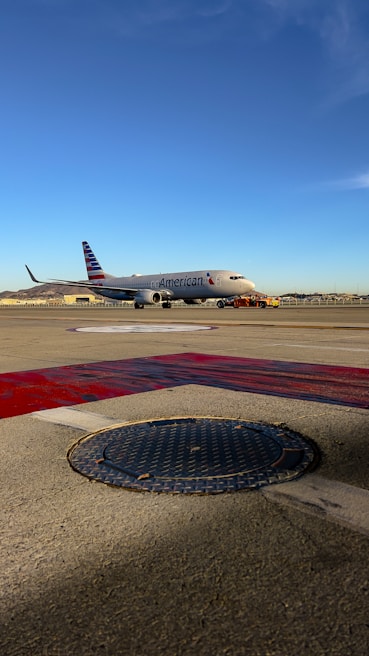 An airplane on the tarmac at an airport with the American Airlines logo. The sky is clear and blue, and there are distant hills and airport structures in the background. The foreground shows a textured manhole cover and painted markings on the ground.