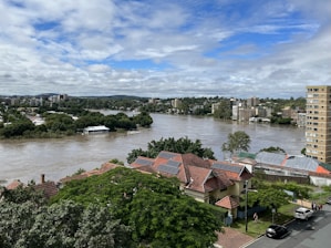 A river flows through an urban area, with trees and residential houses on the banks. The water level appears high, suggesting possible flooding. In the background, there are several multi-story apartment buildings and a partially cloudy sky.