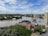 A river flows through an urban area, with trees and residential houses on the banks. The water level appears high, suggesting possible flooding. In the background, there are several multi-story apartment buildings and a partially cloudy sky.