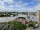 A river flows through an urban area, with trees and residential houses on the banks. The water level appears high, suggesting possible flooding. In the background, there are several multi-story apartment buildings and a partially cloudy sky.
