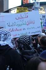 A crowd of people holds signs with various messages, including calls for resistance and liberation. One prominent sign is written in both Arabic and English, focusing on the phrase 'Palestine is ours' and mentions a fight for complete liberation. Many attendees wear patterned headscarves, and there is a camera visible as someone takes photographs of the event.
