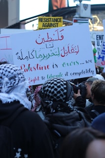 A crowd of people holds signs with various messages, including calls for resistance and liberation. One prominent sign is written in both Arabic and English, focusing on the phrase 'Palestine is ours' and mentions a fight for complete liberation. Many attendees wear patterned headscarves, and there is a camera visible as someone takes photographs of the event.