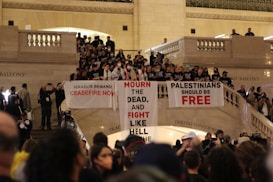 A large group of people gathered in a grand indoor setting, possibly a train station or historical building, holding banners with political messages. The crowd is diverse, with individuals wearing masks and matching shirts. The architecture features marble elements, high ceilings, and visible balconies.