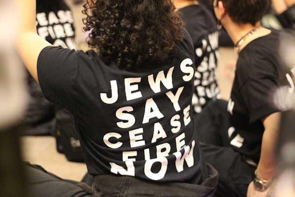 Several people are sitting on the ground wearing black t-shirts with bold white letters that read 'JEWS SAY CEASE FIRE NOW.' The scene captures a peaceful protest or demonstration setting.