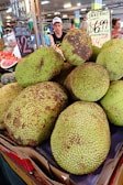 A pile of large jackfruits is displayed at a market stall. The bumpy green fruit has dark patches and is priced at $6.99 per kilo. Various signs surround the display, indicating prices for other fruits like seedless watermelon. People browse the market in the background, creating a bustling atmosphere.