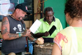 Three individuals are engaged in preparing coconuts. One person, with tattoos and wearing a cap, is holding a cleaver, while another person in a neon green shirt appears to be assisting. The background includes a sign and various items. A third person stands with their back to the camera observing.