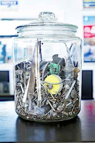 A transparent glass jar filled with assorted metal screws, nails, and bits of hardware. Among the items inside are a circular yellow object, a small green figure, and various metallic and wooden fragments. The jar sits on a dark surface with a blurred background, possibly indicating a workshop or storage environment.