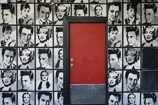 A mural featuring monochromatic portraits of iconic figures covers the wall around a red door. The artwork includes repeated images of famous personalities in a grid-like pattern.