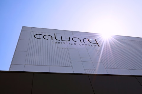 A modern building facade with the text 'Calvary Christian Church' prominently displayed. The structure features a minimalist design with clean lines and a sunburst effect in the top right corner, suggesting a sunny day.
