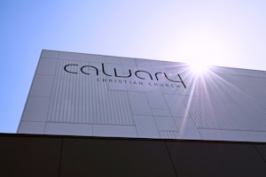A modern building facade with the text 'Calvary Christian Church' prominently displayed. The structure features a minimalist design with clean lines and a sunburst effect in the top right corner, suggesting a sunny day.