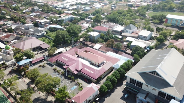 A residential area with a mixture of buildings, trees, and roads viewed from above. The central focus is on a distinct building with red roofs labeled 'PANGKEP' and 'PAREES'. There are basketball courts, other smaller structures, and a large building with a grey roof on the right.