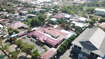A residential area with a mixture of buildings, trees, and roads viewed from above. The central focus is on a distinct building with red roofs labeled 'PANGKEP' and 'PAREES'. There are basketball courts, other smaller structures, and a large building with a grey roof on the right.