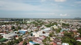 Aerial view of a town with a mixture of residential buildings, commercial structures, and greenery. Mosques and other tall buildings can be seen in various parts of the town. The town is surrounded by a vast landscape that stretches out to the horizon.
