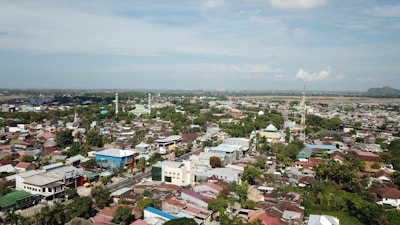 Aerial view of a town with a mixture of residential buildings, commercial structures, and greenery. Mosques and other tall buildings can be seen in various parts of the town. The town is surrounded by a vast landscape that stretches out to the horizon.