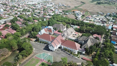 An aerial view of a densely populated urban area featuring a cluster of buildings with red and grayish roofs surrounded by lush greenery. The scene includes a mix of residential and possibly institutional structures with a winding road running through the area. In the foreground, there's a multi-use open space, possibly a court, adjacent to a tree-lined street. The background features agricultural fields stretching toward the horizon.