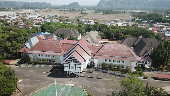 A large complex with multiple buildings featuring red roofs and white walls, surrounded by a lush landscape and situated adjacent to a town. The complex appears to be an institutional or governmental building with a well-maintained exterior. In the background, there are residential houses, trees, and mountains.