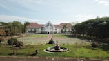 A large building with white walls and a red roof sits in the background, surrounded by a wide open courtyard and lush green lawns. In the foreground, there's a circular fountain with several tiers. The area is enclosed by numerous trees and vegetation, providing a serene and orderly environment.