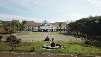 A large building with white walls and a red roof sits in the background, surrounded by a wide open courtyard and lush green lawns. In the foreground, there's a circular fountain with several tiers. The area is enclosed by numerous trees and vegetation, providing a serene and orderly environment.