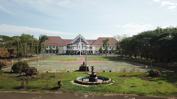 A large building with white walls and a red roof sits in the background, surrounded by a wide open courtyard and lush green lawns. In the foreground, there's a circular fountain with several tiers. The area is enclosed by numerous trees and vegetation, providing a serene and orderly environment.