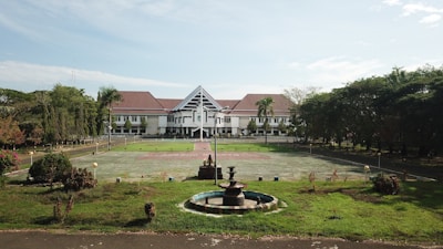 A large building with white walls and a red roof sits in the background, surrounded by a wide open courtyard and lush green lawns. In the foreground, there's a circular fountain with several tiers. The area is enclosed by numerous trees and vegetation, providing a serene and orderly environment.