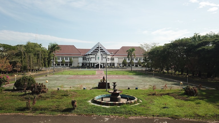 A large building with white walls and a red roof sits in the background, surrounded by a wide open courtyard and lush green lawns. In the foreground, there's a circular fountain with several tiers. The area is enclosed by numerous trees and vegetation, providing a serene and orderly environment.