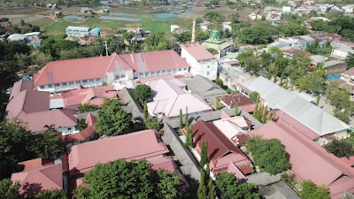 Aerial view of a sprawling complex of buildings with red roofs, surrounded by lush greenery and trees. The area includes a mosque with a distinctive green dome and minaret, situated in a small town with other residential and commercial buildings scattered around.