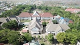 An aerial view captures a complex of buildings with gabled roofs surrounded by lush greenery and trees. The structures are arranged in a U-shape with a central courtyard, and some rooftops are covered with blue and red tiles. Behind the buildings, there are expansive fields and residential areas stretching to the horizon.