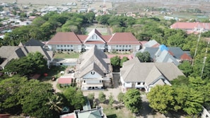 An aerial view captures a complex of buildings with gabled roofs surrounded by lush greenery and trees. The structures are arranged in a U-shape with a central courtyard, and some rooftops are covered with blue and red tiles. Behind the buildings, there are expansive fields and residential areas stretching to the horizon.