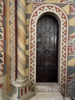 An ornately decorated medieval-style door with iron embellishments set in a vibrantly painted wall. The wall features intricate geometric and floral patterns in earthy tones of red, yellow, and grey, with a spiral column adjacent to the door.