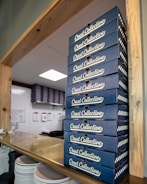 A tall stack of blue boxes labeled 'Crust Collective' is neatly arranged on a wooden counter. In the background, additional stacks are visible against a white, organized kitchen wall with clipboards hanging. White plates are stacked at the bottom left corner.