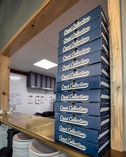 A tall stack of blue boxes labeled 'Crust Collective' is neatly arranged on a wooden counter. In the background, additional stacks are visible against a white, organized kitchen wall with clipboards hanging. White plates are stacked at the bottom left corner.