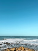 A serene ocean scene with gentle waves crashing onto a rocky shoreline under a clear blue sky. The water's surface reflects different shades of blue, creating a calming and peaceful atmosphere.