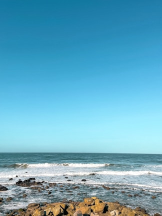 A serene ocean scene with gentle waves crashing onto a rocky shoreline under a clear blue sky. The water's surface reflects different shades of blue, creating a calming and peaceful atmosphere.