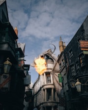 A large, realistic dragon is perched atop a building, breathing fire against a backdrop of old-fashioned, slightly darkened architecture. The scene is set in a narrow street filled with vintage-style shops and lanterns, with a cloudy sky overhead.