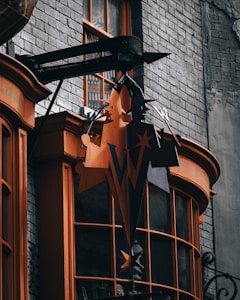 A store sign with a distinctive 'W' logo, featuring stars and a whimsical design, hangs in front of a brick building. The sign and window frames are painted in a vibrant orange color, contrasting with the gray brick background. The building appears slightly aged, adding a vintage feel.
