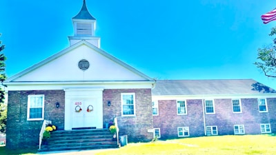A brick building with a white steeple and gabled roof is situated in a grassy area. The entrance features double doors decorated with wreaths and the number 455 above. Windows are symmetrically placed along the building's facade, and an American flag is partially visible on the right. Yellow flowers in planters flank the steps leading to the entrance.