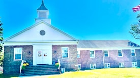 A brick building with a white steeple and gabled roof is situated in a grassy area. The entrance features double doors decorated with wreaths and the number 455 above. Windows are symmetrically placed along the building's facade, and an American flag is partially visible on the right. Yellow flowers in planters flank the steps leading to the entrance.