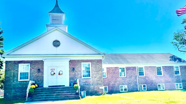 A brick building with a white steeple and gabled roof is situated in a grassy area. The entrance features double doors decorated with wreaths and the number 455 above. Windows are symmetrically placed along the building's facade, and an American flag is partially visible on the right. Yellow flowers in planters flank the steps leading to the entrance.