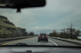 A road trip scene with a view through a car's windshield of a highway. The road is flanked by guardrails and bordered by rocky hills on the left and sparse trees on the right. A car is driving ahead, and a snow-capped mountain stands in the background under an overcast sky.