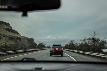 A road trip scene with a view through a car's windshield of a highway. The road is flanked by guardrails and bordered by rocky hills on the left and sparse trees on the right. A car is driving ahead, and a snow-capped mountain stands in the background under an overcast sky.