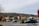 A commercial area with various stores like Wells Fargo, Dollar Tree, and Rivermark Credit Union. A blue SUV is parked in the lot, and a 'For Lease' sign is visible. The background shows a hill with scattered vegetation under a cloudy sky. A leafless tree is on the left.