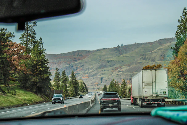 A wide shot of a car carrier navigating through a scenic highway surrounded by green trees.