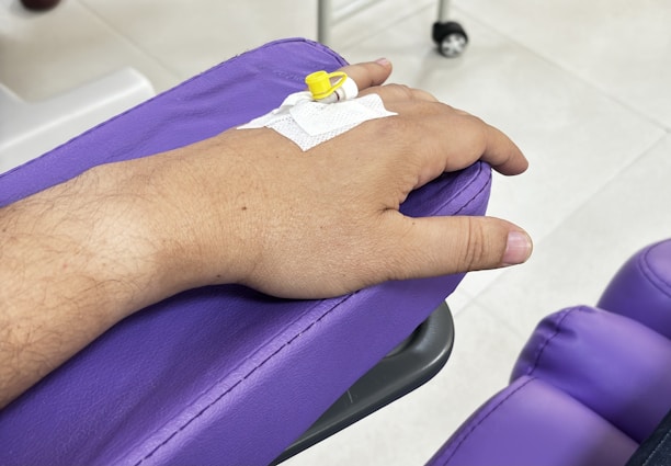 A hand rests on a purple cushion, connected to an intravenous line with a yellow cap. The background includes a tiled floor and a metallic stand, possibly part of medical equipment.