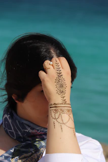 A smiling teen wearing a henna-adorned hand and layered bracelets against a sunset beach scene.