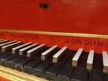 A detailed view of a keyboard from a musical instrument, with black and white keys. The background is bright red with gold trim, featuring the words 'Bavington' and 'London' in black lettering.