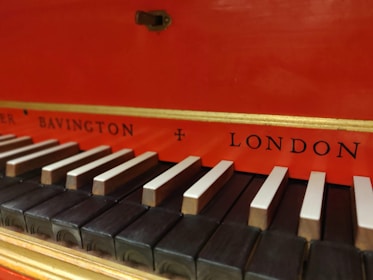 A detailed view of a keyboard from a musical instrument, with black and white keys. The background is bright red with gold trim, featuring the words 'Bavington' and 'London' in black lettering.