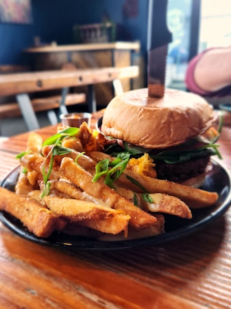 A close-up of a savory meal featuring a juicy burger with fresh greens, cheese, and a bun, with a knife inserted into the top. Crispy, seasoned fries are served on the side, garnished with chopped green onions. The setting is a rustic wooden table in a cozy dining area.